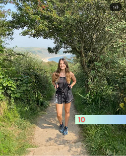 A white female with long brown hair smiles at the camera on a coastal path. 